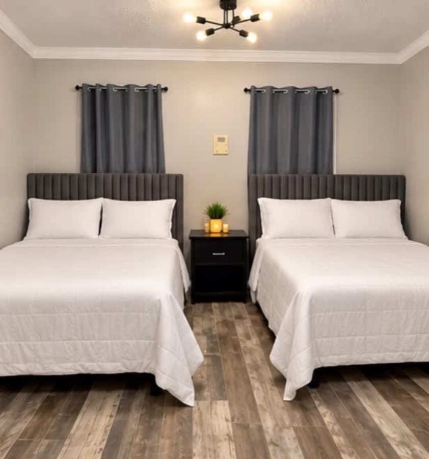 A bedroom with two beds, grey channel-tufted headboards, white bedding and a shared nightstand on wood-look flooring.