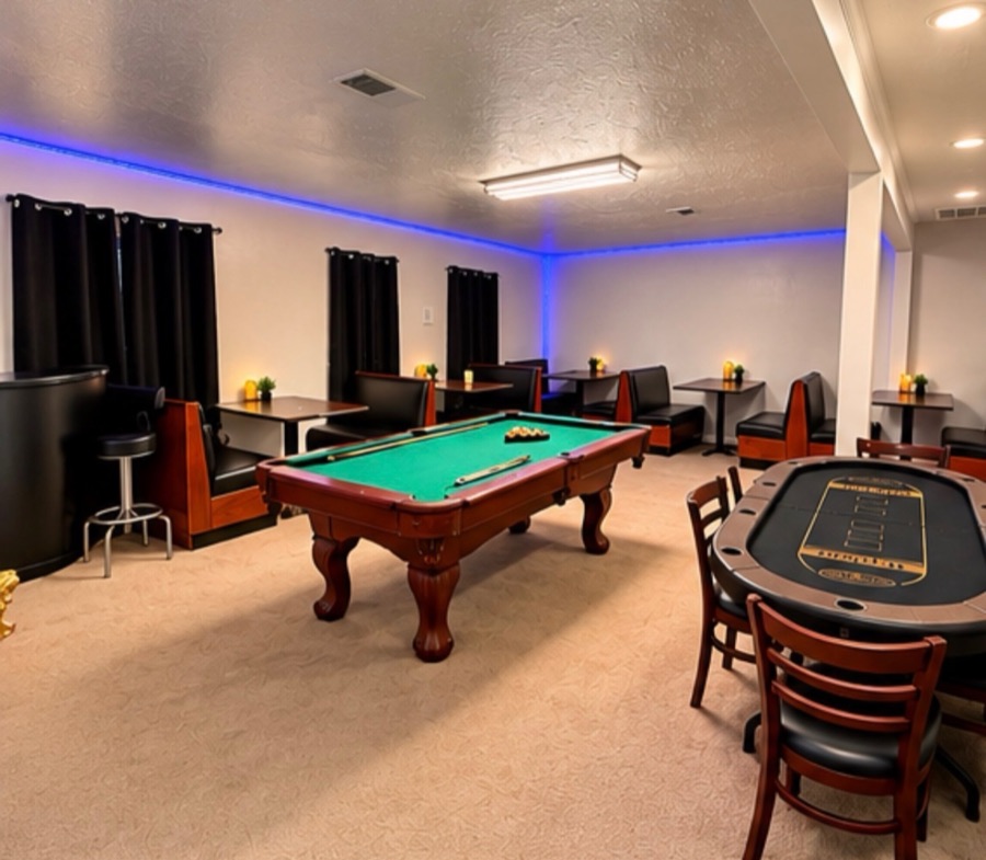 The game room: a pool table with cues laid across the felt, a poker table with chairs, and booth seating under blue cove lighting.
