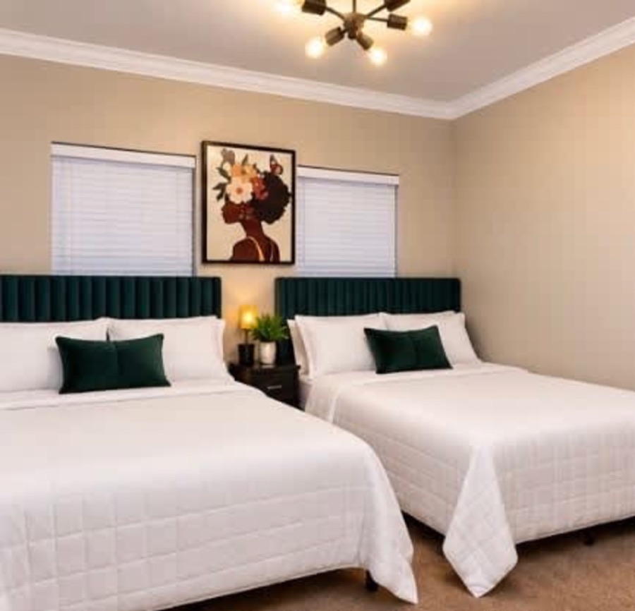A bedroom with two beds, green upholstered headboards, white bedding and a framed portrait on the wall between the windows.
