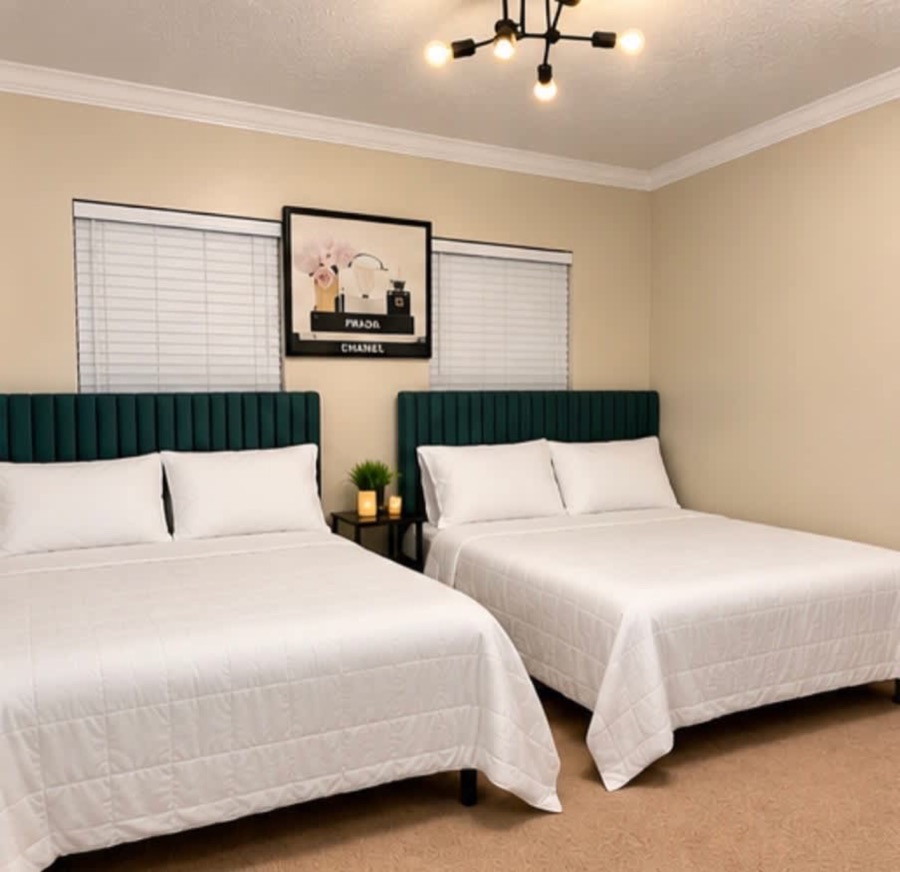 A bedroom with two beds, deep green channel-tufted headboards and white bedding, framed art between two blinded windows.