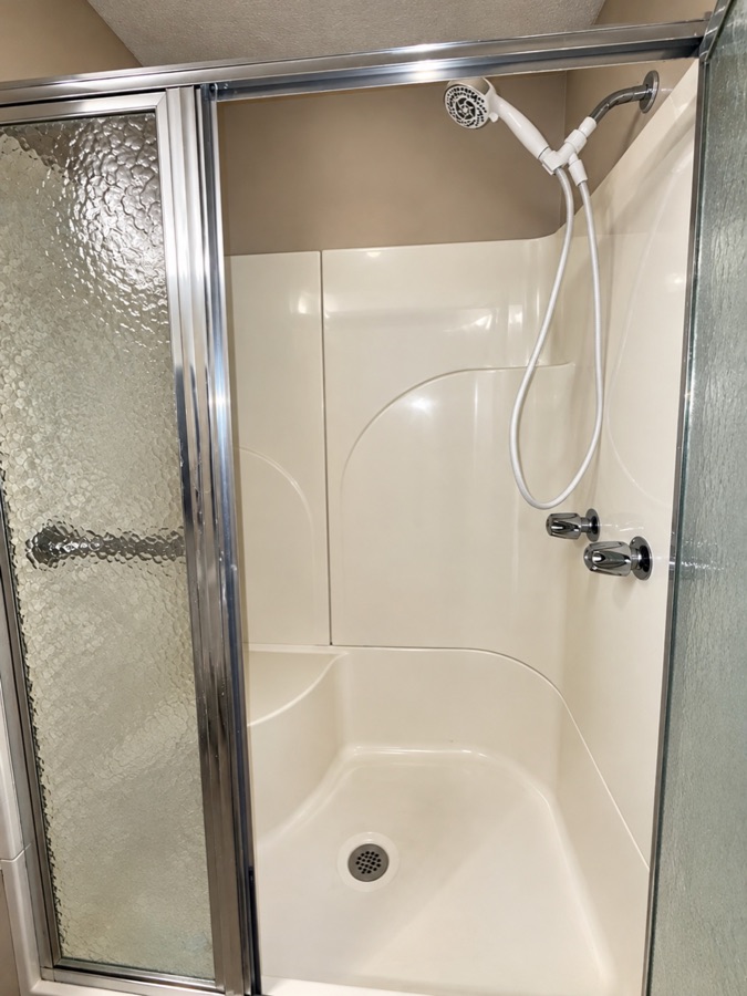 A white walk-in shower stall with a chrome-framed glass door and a handheld shower head.
