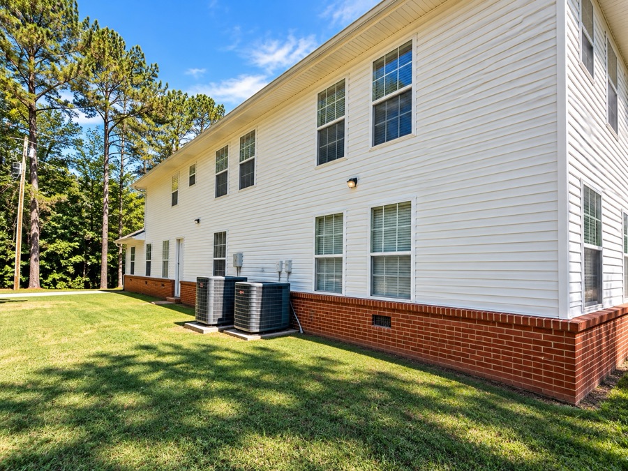The side of the house in daylight: white siding above a red brick base, with pines and open lawn beyond.