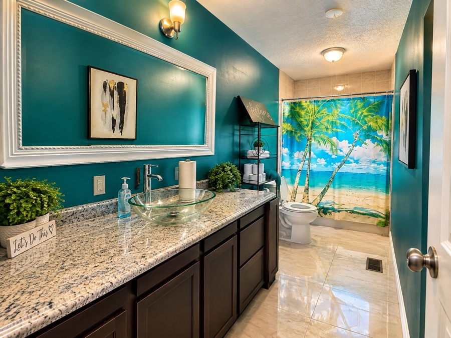 A bathroom with deep teal walls, a long granite counter with a glass vessel sink, a wide white-framed mirror and a palm-print shower curtain.