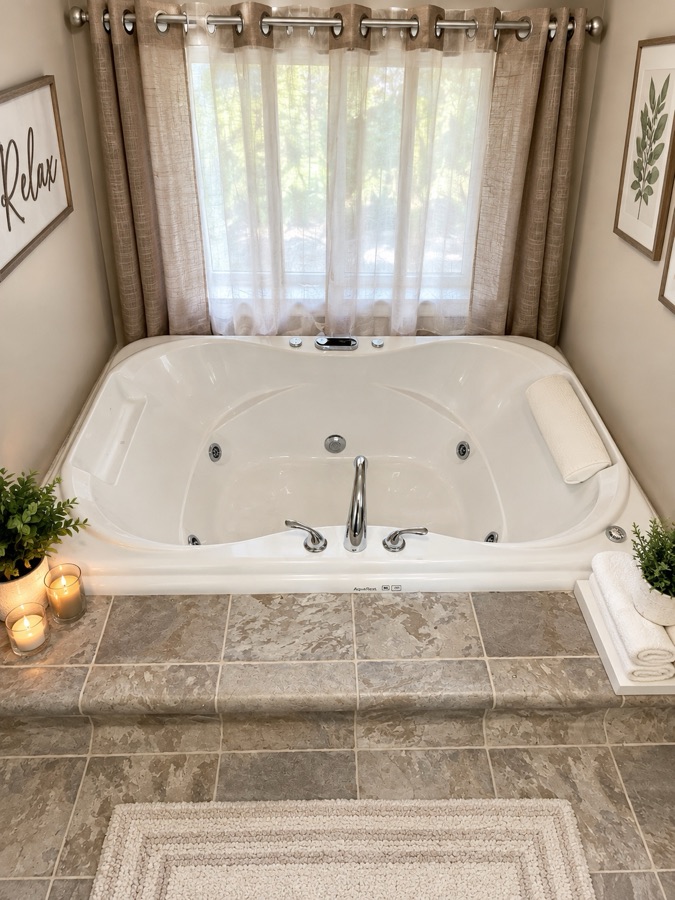 The jetted tub set into a tiled surround beneath a curtained window, with lit candles and rolled white towels on the ledge beside it.